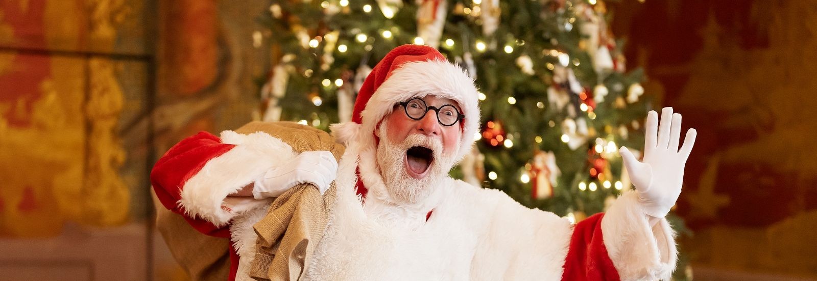 A cheerful Santa slings a sack of presents over his shoulder with an open mouthed expression. Behind him is a grand, tall Christmas tree well decorated with ornaments and fairy lights.