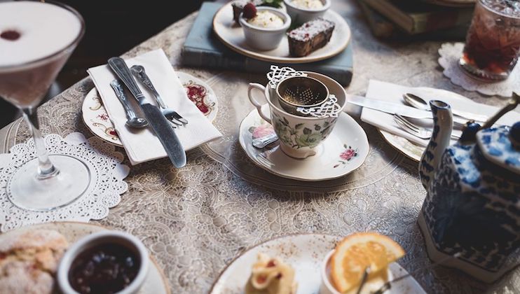 A table set for afternoon tea, featuring a teacup with a strainer, a teapot, cutlery on napkins, a lace tablecloth, plates of assorted desserts, and a glass with a pink drink. The setting is decorated with elegant and vintage-style items.