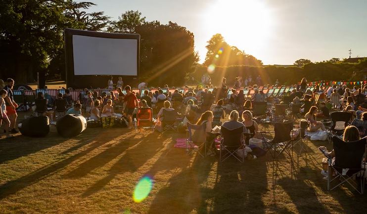 A large group of people sitting on the grass and in chairs at an outdoor event during sunset. They are gathered in front of a big screen, possibly for an open-air movie screening. Colorful pennant banners decorate the area, and long shadows are cast by the setting sun.