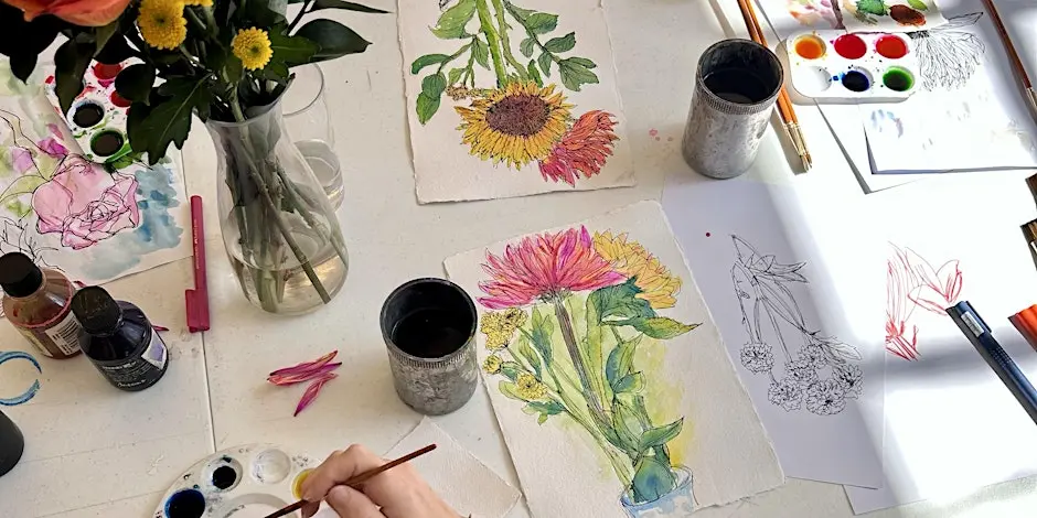 A desktop shot of watercolour study painting. Vase of flowers as reference, a hand with a paintbrush dips into wet paint. The desk is scattered with pens, brushes, pallettes of colourful paint, and gorgeous watercolour studies of the flowers.