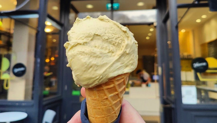 A hand holds up a cone with a large scoop of pistachio ice cream in front of a cafe entrance. The background shows the open doorway of the cafe with people inside and tables with chairs outside. The ice cream is light green and appears creamy.