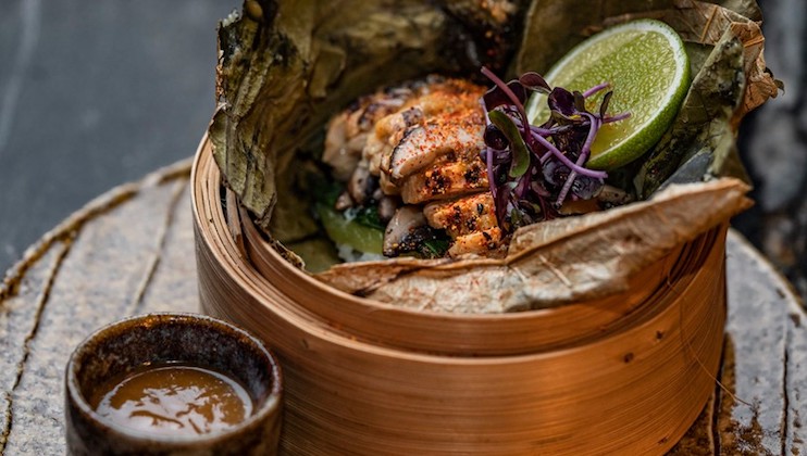 A close-up of a dish served in a bamboo steamer. It features grilled meat topped with microgreens, garnished with lime slices, and encased in a leaf. A small bowl of dipping sauce is placed beside the steamer on a rustic surface.