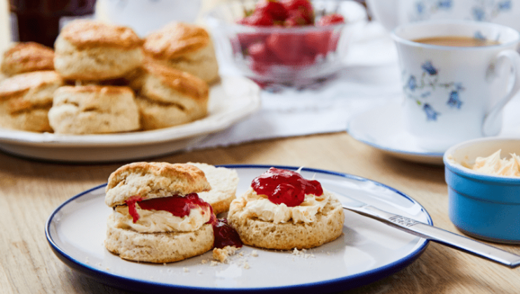 A plate with a scone topped with clotted cream and strawberry jam, a knife on the side. In the background, a bowl of more strawberry jam, a plate with more scones, and a cup of tea in a floral teacup are visible. The scene is set on a wooden table.