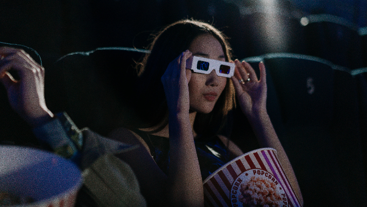 A person with long hair is sitting in a dark movie theater wearing 3D glasses. The person is holding a bucket of popcorn and has one hand adjusting their glasses. Empty seats are visible in the background.