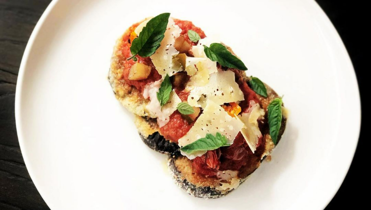 A white plate holds a piece of bruschetta topped with tomato sauce, grated Parmesan cheese, basil leaves, black pepper, and small tomato pieces. The dish appears to be garnished with herbs and is set against a dark background.