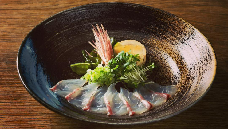 A plated dish features thinly sliced sashimi with garnishes of microgreens, scallions, and a piece of seafood topped with delicate threads, artfully arranged in a dark, textured bowl on a wooden surface.