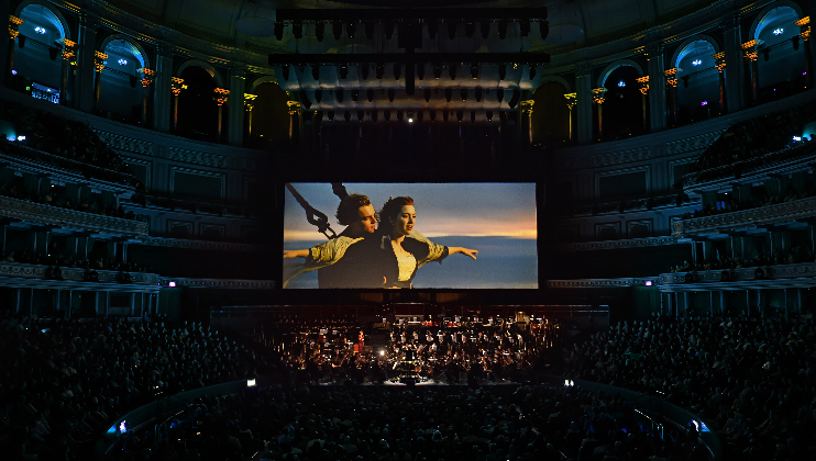 A large orchestra performs in a grand concert hall with an audience watching a giant screen above the stage. The screen displays a famous scene from a movie, where a couple stands at the bow of a ship with open arms against a sunset-lit ocean backdrop.
