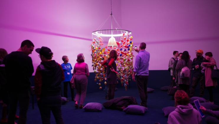 A group of people stand around an artistic installation featuring a suspended circular frame with colorful hanging objects, illuminated by purple lighting. The room has pillows scattered on the floor, and attendees are observing and interacting with the artwork.