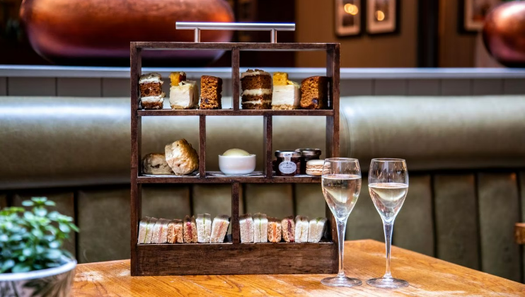 A wooden multi-tiered stand is filled with various pastries, cakes, scones, and sandwiches. The stand is set on a wooden table alongside two glasses of white wine. A small plant is also on the table. The background features a cushioned bench and framed pictures on the wall.