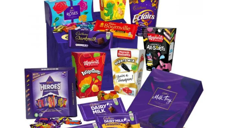 An assortment of colorful Cadbury chocolate and candy boxes is displayed. Products include Dairy Milk, Roses, Bournville, Éclairs, Wine Gums, Liquorice Allsorts, Milk Tray, and Heroes. Individual chocolates and candies are also visible, arranged randomly around the boxes.
