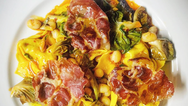 A plate of pasta featuring wide, flat noodles topped with slices of cured meat, beans, leafy greens, and vegetables. The dish is garnished with a light sauce, and the ingredients are vibrant and colorful, creating an appetizing presentation.
