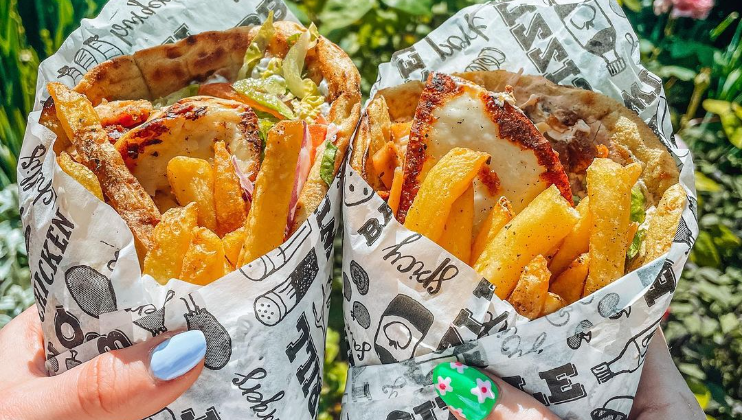 Two hands holding wrapped gyros filled with meat, fries, lettuce, and other ingredients. The wraps are covered in white paper with various black illustrations and text. Lush greenery is visible in the blurred background. One hand has blue nail polish and a floral nail design.