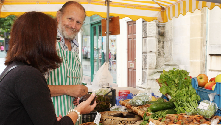 A vendor with a striped apron smiles while serving a customer at an outdoor market stall. The table is laden with fresh produce, including cucumbers, apples, carrots, and leafy greens. The customer, wearing glasses and a dark sweater, examines a vegetable.