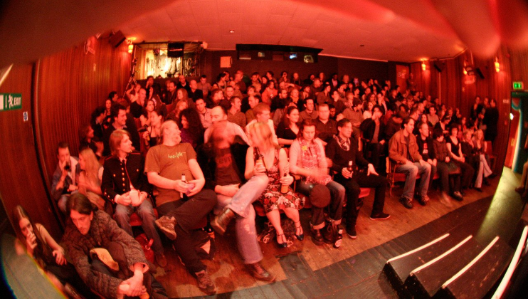A dimly lit, crowded venue with rows of seated people facing a stage. The audience appears engaged, with some individuals chatting and others looking towards the front. The room has a warm ambiance with wooden elements and exit signs visible.