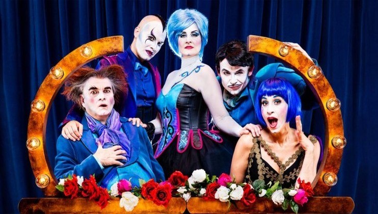 A group of five dramatically costumed performers pose in front of a blue curtain. The central figure has blue hair and a black corset dress. Four others surrounding her have exaggerated makeup and vibrant costumes, all framed by a semicircle of lights and red roses.