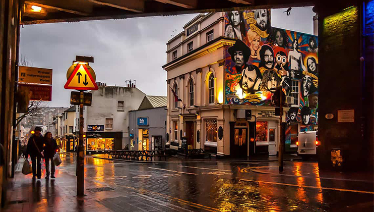 A rainy street scene at dusk with two people walking under an umbrella. Buildings are illuminated in the background, including one with a large, colorful mural featuring various faces. Reflections from the wet pavement add to the vibrant atmosphere.