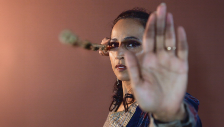 Urja Desai Thakore poses with her left hand extended outward (covering half her face); with her right hand she points a stick toward the viewer