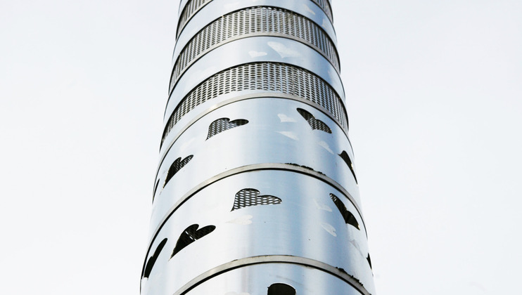 A tall metal structure with circular segments, featuring heart-shaped cutouts and mesh patterns. The background is a pale, overcast sky.