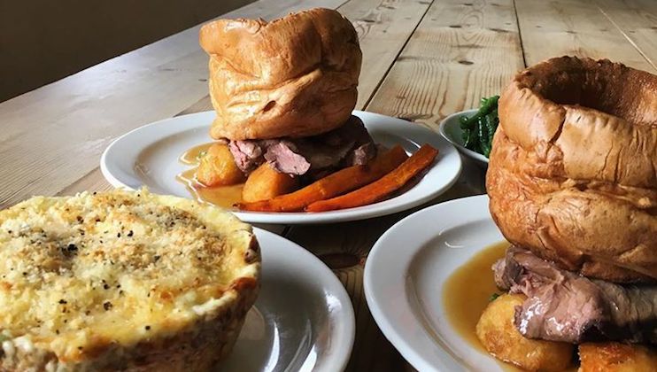 Two plates with roast beef and Yorkshire pudding, served with roasted carrots and potatoes, are placed on a wooden table. Next to them is a dish of broccoli and a pie covered with melted cheese on a separate plate.