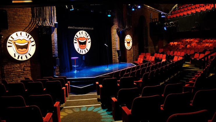 A dimly lit comedy club with a stage set with a microphone on a stand and a stool. The room features red theater seating and three circular logos with a laughing mouth design. The background includes a brick wall and a website URL displayed on a banner.