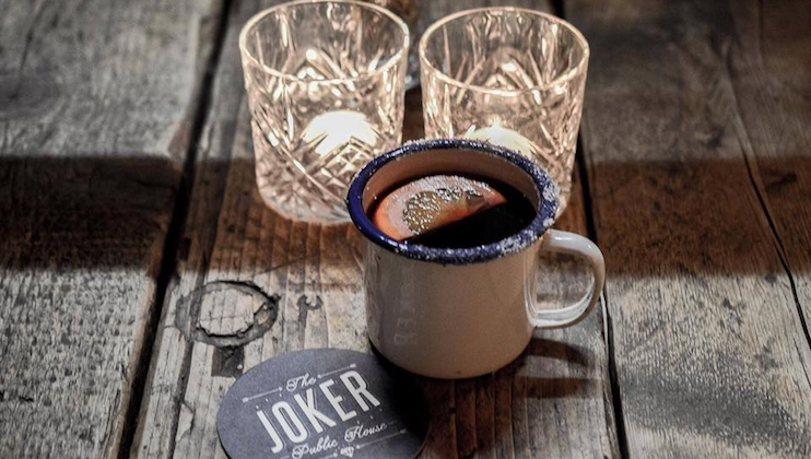 A cup filled with a dark beverage, garnished with a slice of orange and placed on a wooden table next to two empty, textured glass candle holders. A coaster that reads The Joker Public House is placed near the cup.