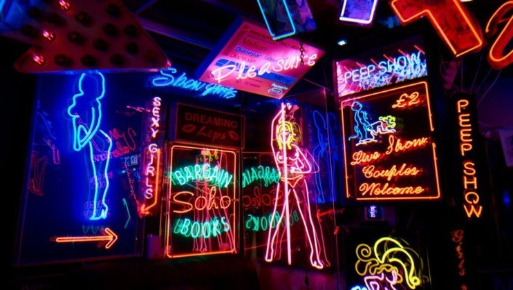 The image features brightly colored neon signs in various shapes and sizes, advertising adult entertainment. Words like Pleasure, Sexy Girls, Peep Show, and Live Show Couples Welcome are prominently displayed alongside neon silhouettes of women.