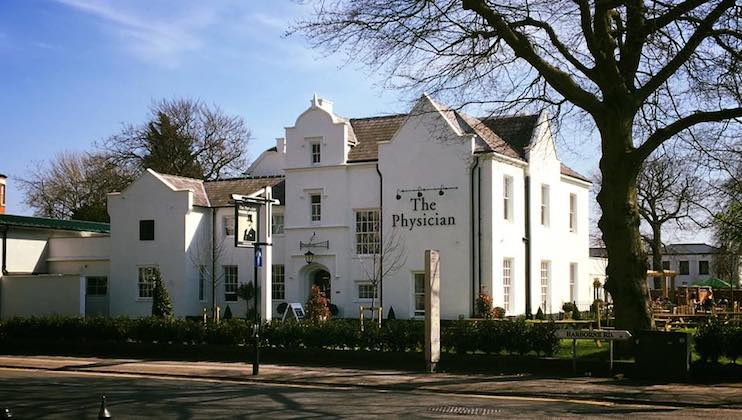 A large white building with a classic architectural style houses The Physician, a pub and restaurant. The establishment is surrounded by trees and outdoor seating areas. A sign and lighting fixtures adorn the exterior. The sky is clear and sunny.