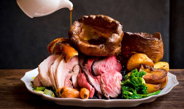 A dish featuring slices of roasted meat, crispy roast potatoes, Yorkshire puddings, carrots, and green vegetables. Gravy is being poured over the meal from a white jug above the plate. The meal is arranged on a white plate against a dark background.
