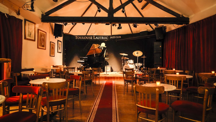 The interior of Toulouse Lautrec jazz club's third floor; around twenty tables are arranged before the stage (on which there is a piano and microphone)