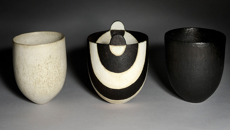 Three ceramic vessels displayed side by side. The left one is a smooth, white cup, the middle vessel features a combination of white and black geometric patterns with a circular design at the top, and the right vessel is entirely black with a textured surface.