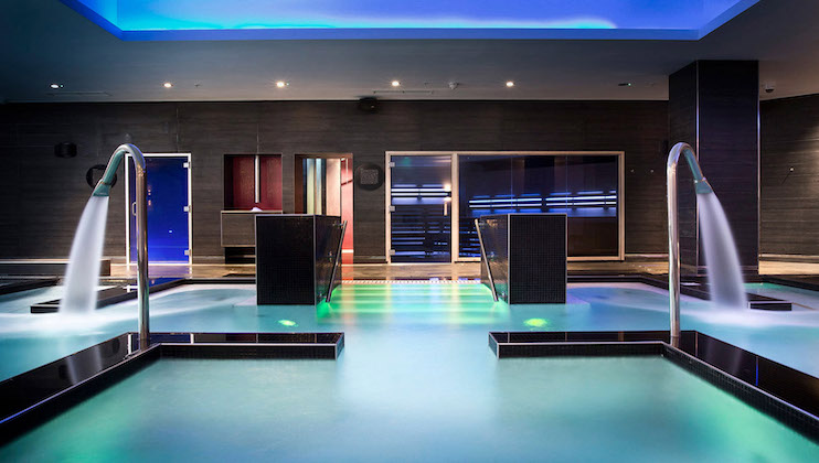 Luxurious indoor pool area with sleek, modern design. Four water spouts gently cascade into the pool. Ambient lighting creates a serene environment, with blue hues reflecting off the water. Pathway in the middle and futuristic wall and ceiling accents enhance the ambiance.