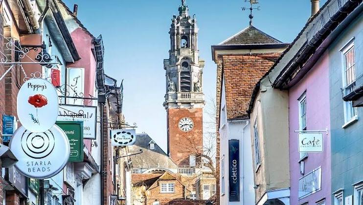 A bustling street featuring colorful buildings lined with various shops and signs, including Poppy's and Starla Beads. In the background, a prominent church tower with a clock stands tall against a clear blue sky.