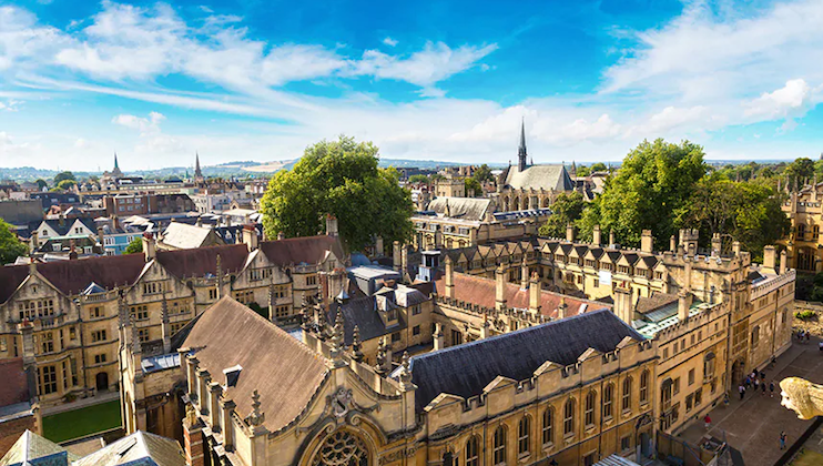 A panoramic view of an ancient city featuring numerous historic buildings with tall spires, ornate architecture, and lush green trees. The sky is bright blue with scattered white clouds, enhancing the overall picturesque scene.