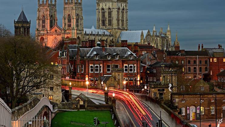Night view of a historic cityscape featuring illuminated buildings and York Minster Cathedral with light trails from vehicles on a busy street in the foreground. The sky is overcast, adding a dramatic effect to the scene.