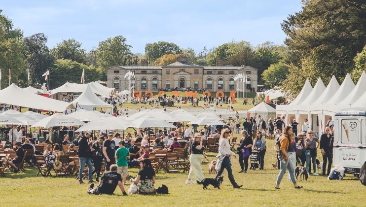 A large outdoor festival with numerous white tents and people scattered across a grassy area. Attendees are walking, sitting on blankets, and socializing. In the background, a grand building is visible. The weather is clear with a blue sky and trees surrounding the area.
