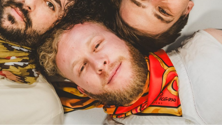 Sol Chase, in a white suit and red-orange cravatte, smiles at the camera above him; he is lying with his head in formation with the other members of Big Love Car Wash