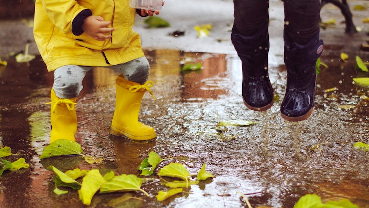 Two children play in a puddle on a rainy day. One wears a yellow raincoat and matching rain boots while the other has black rain boots. They joyfully splash water, surrounded by wet, fallen leaves scattered on the pavement. Only their lower bodies are visible.