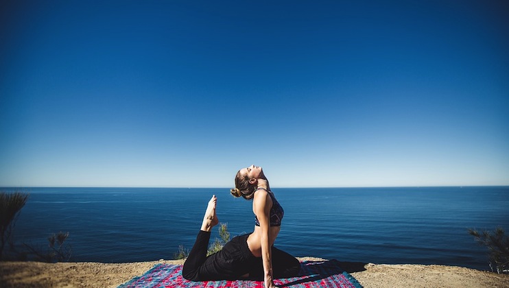 A person practices yoga on a colorful mat at a cliffside overlooking a calm, expansive ocean and a clear blue sky. They are performing a backbend pose and appear focused and serene. Sparse vegetation is visible around the cliff's edge.