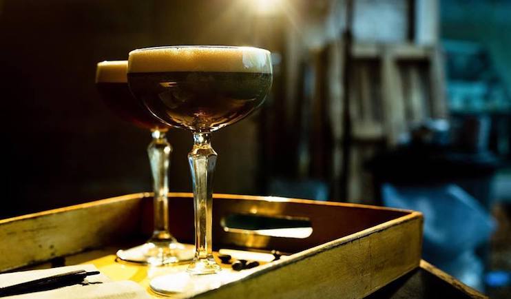 Two elegant cocktail glasses filled with a dark, frothy drink rest on a rustic wooden tray. The background appears blurred with a soft, warm light illuminating the glasses, adding a cozy ambiance to the scene.