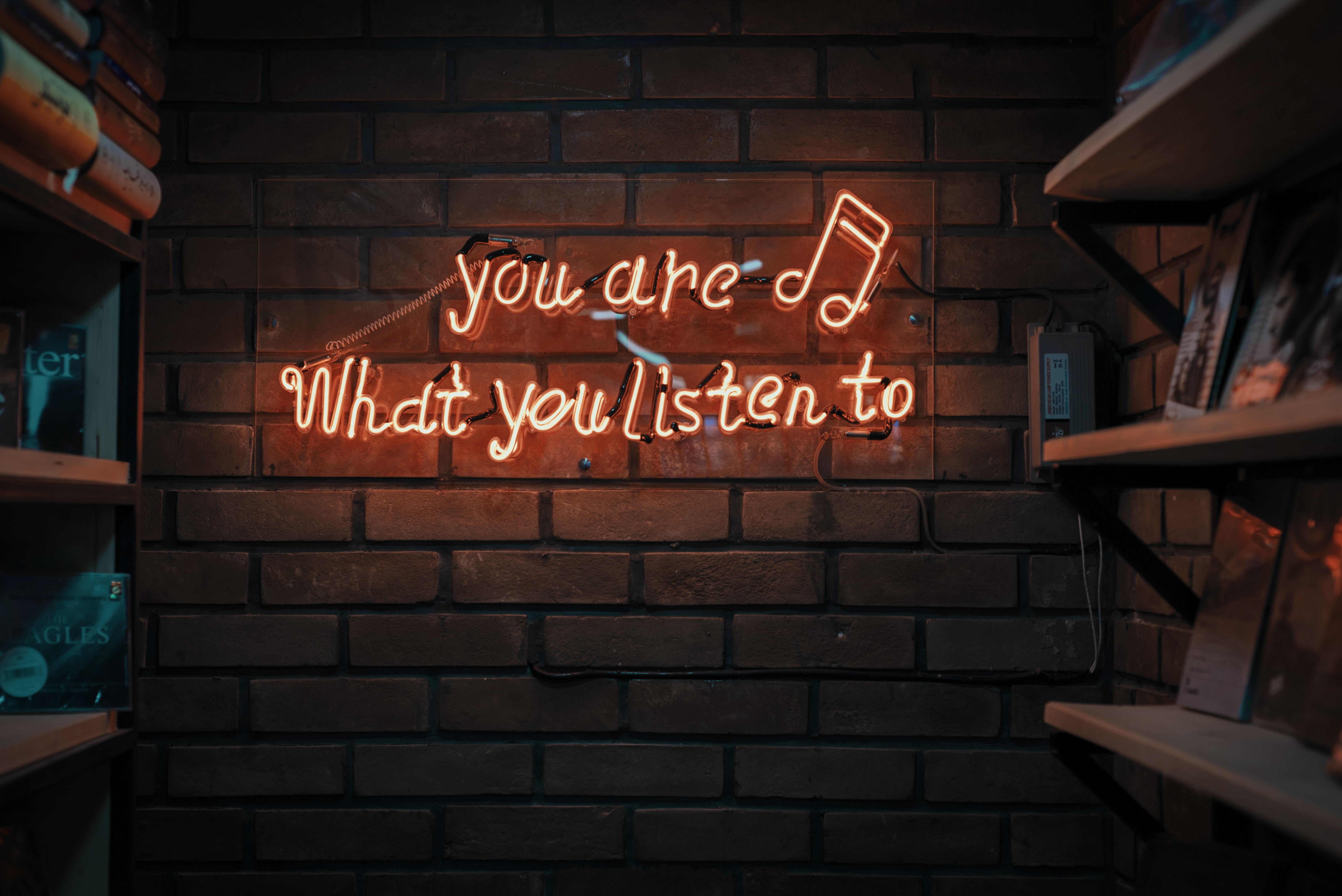 A glowing orange neon sign against a dark brick wall reads you are what you listen to with a musical note symbol. Shelves filled with various books frame the sign on the left and right sides of the image.