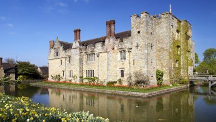 A historic stone castle with crenellated towers and chimneys, surrounded by a moat reflecting the structure. The castle is adorned with red flowers along the perimeter, and lush green vegetation is visible in the background under a clear blue sky.