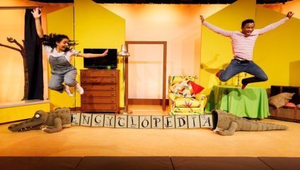 Two children are joyfully jumping in the air in a colorful room decorated like a playful library or reading area. Below them is a toy alligator with blocks spelling ENCYCLOPEDIA. The setting includes bookshelves, a lamp, and vibrant furniture.