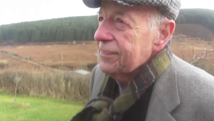 Writer Christopher Burn, author of Poetry Changes Lives, is wrapped up warm and wearing as flat cap as he looks wistfully into the distance in a lush green countryside setting