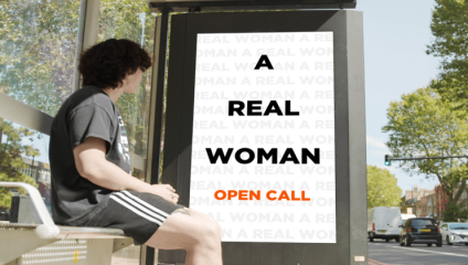 A person with dark curly hair sits at a bus stop, looking at an advertisement. The ad features the text A REAL WOMAN in bold letters, with OPEN CALL at the bottom in orange. The background shows trees and a street scene with parked vehicles.