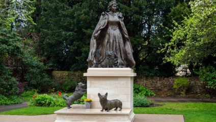 A bronze statue of Queen Elizabeth II with two corgis is situated on a stone pedestal in a garden. The queen is depicted in a regal pose with flowing robes. Greenery and trees surround the setting. A plaque on the pedestal commemorates the years 1926-2022.