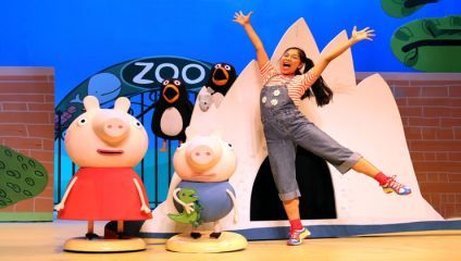 A cheerful performer in colorful clothing and rainbow shoes enthusiastically poses with arms and one leg raised next to two costumed characters: a pink pig in a red dress and another pink pig holding a green dinosaur. The background features a zoo theme.
