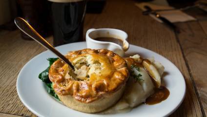 Photograph of a plate of a savoury pie on a bed of mash with gravy and greens