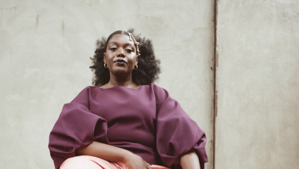 A person with curly hair, wearing a plum-colored top with voluminous sleeves and light pink pants, sits confidently against a textured, neutral-colored wall. They have a calm and poised expression.