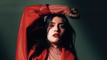 A person with long dark hair, wearing a red jacket over a striped top, poses with their left arm raised and bent at the elbow, casting a shadow. The background is dark and the lighting creates a dramatic effect on their face.