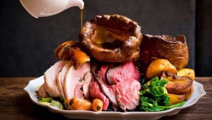 A dish featuring slices of roasted meat, crispy roast potatoes, Yorkshire puddings, carrots, and green vegetables. Gravy is being poured over the meal from a white jug above the plate. The meal is arranged on a white plate against a dark background.